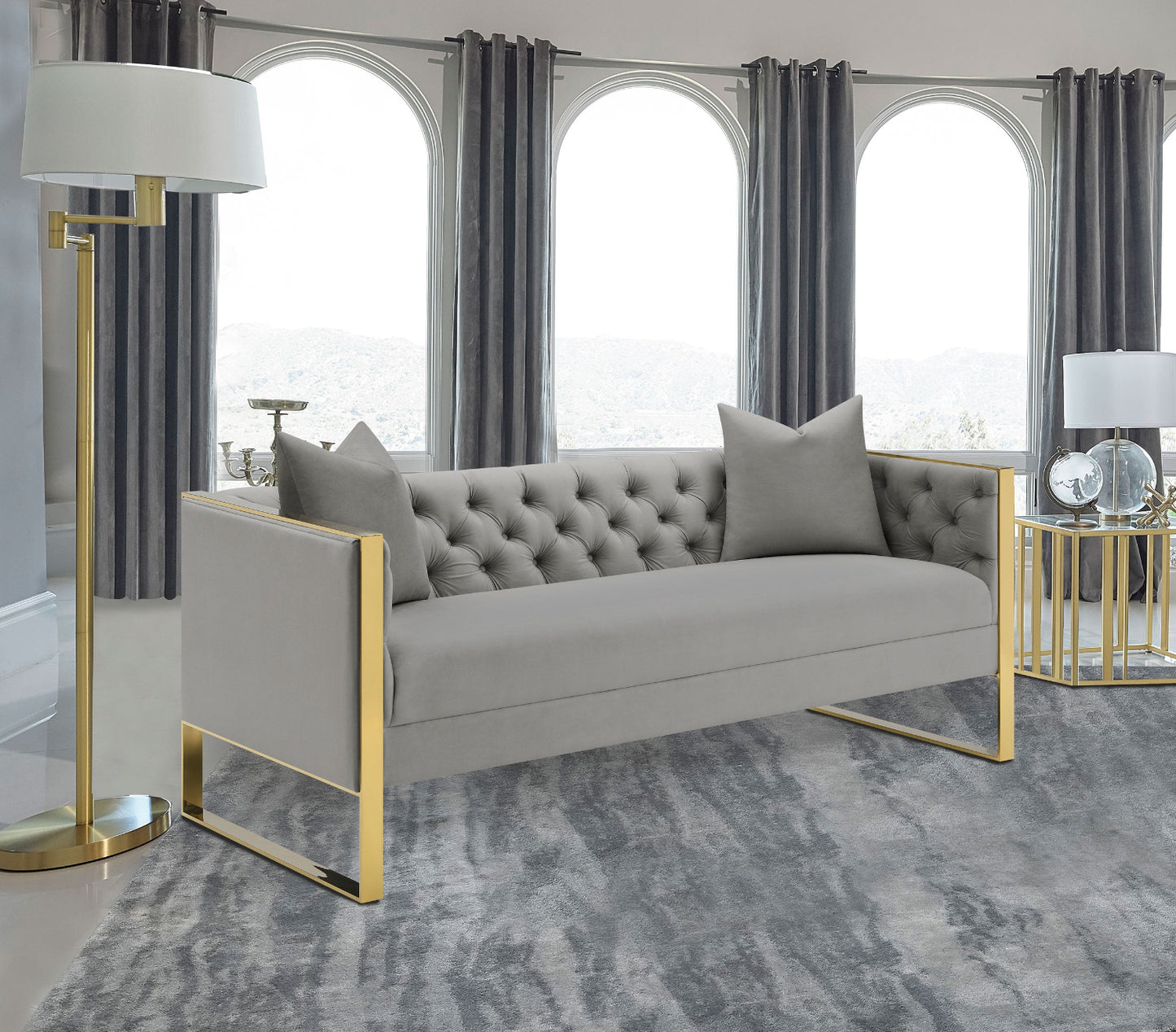 Gray tufted sofa with gold legs in a living room setting with large windows and curtains.