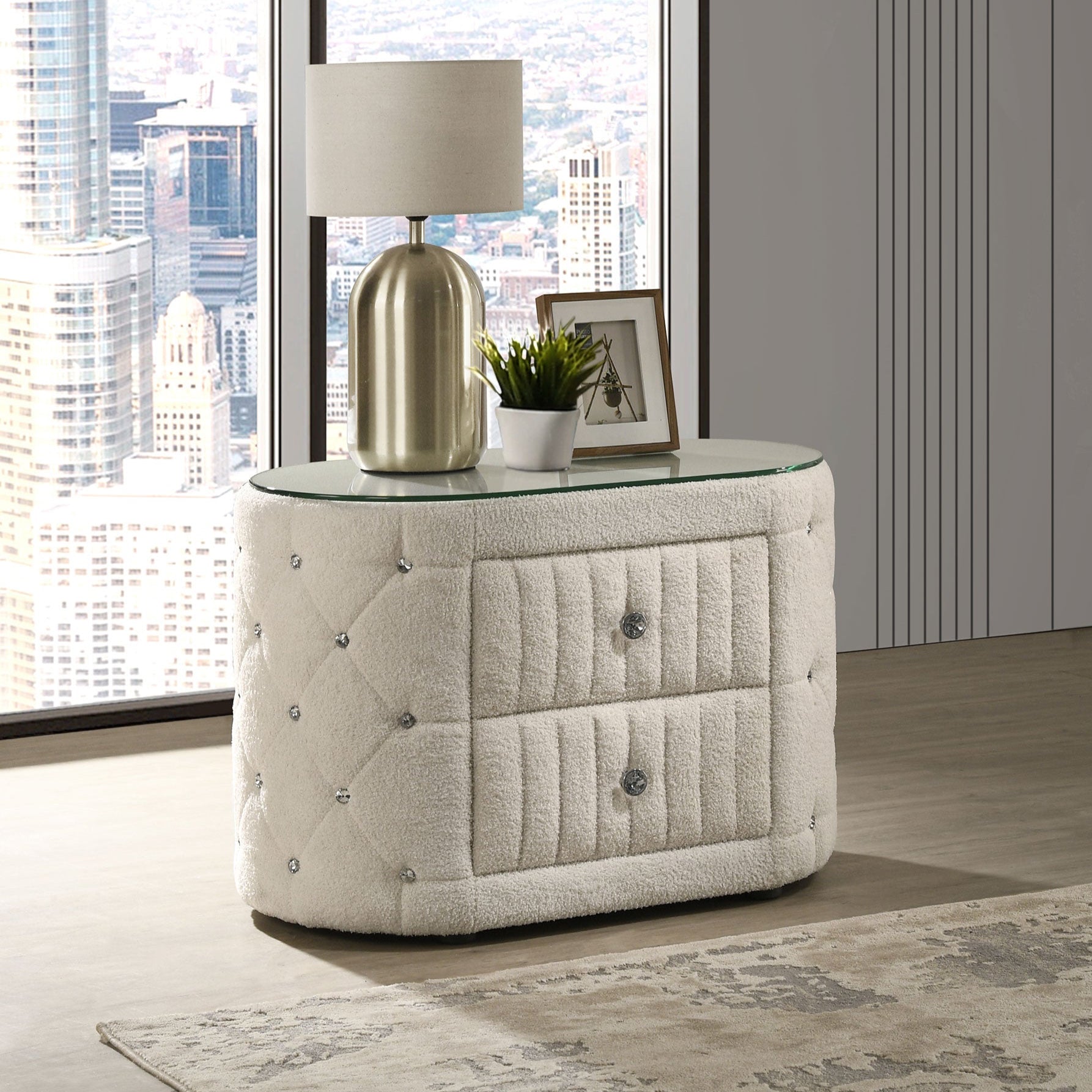 Modern nightstand with lamp and decor in a room with large windows showing a cityscape.